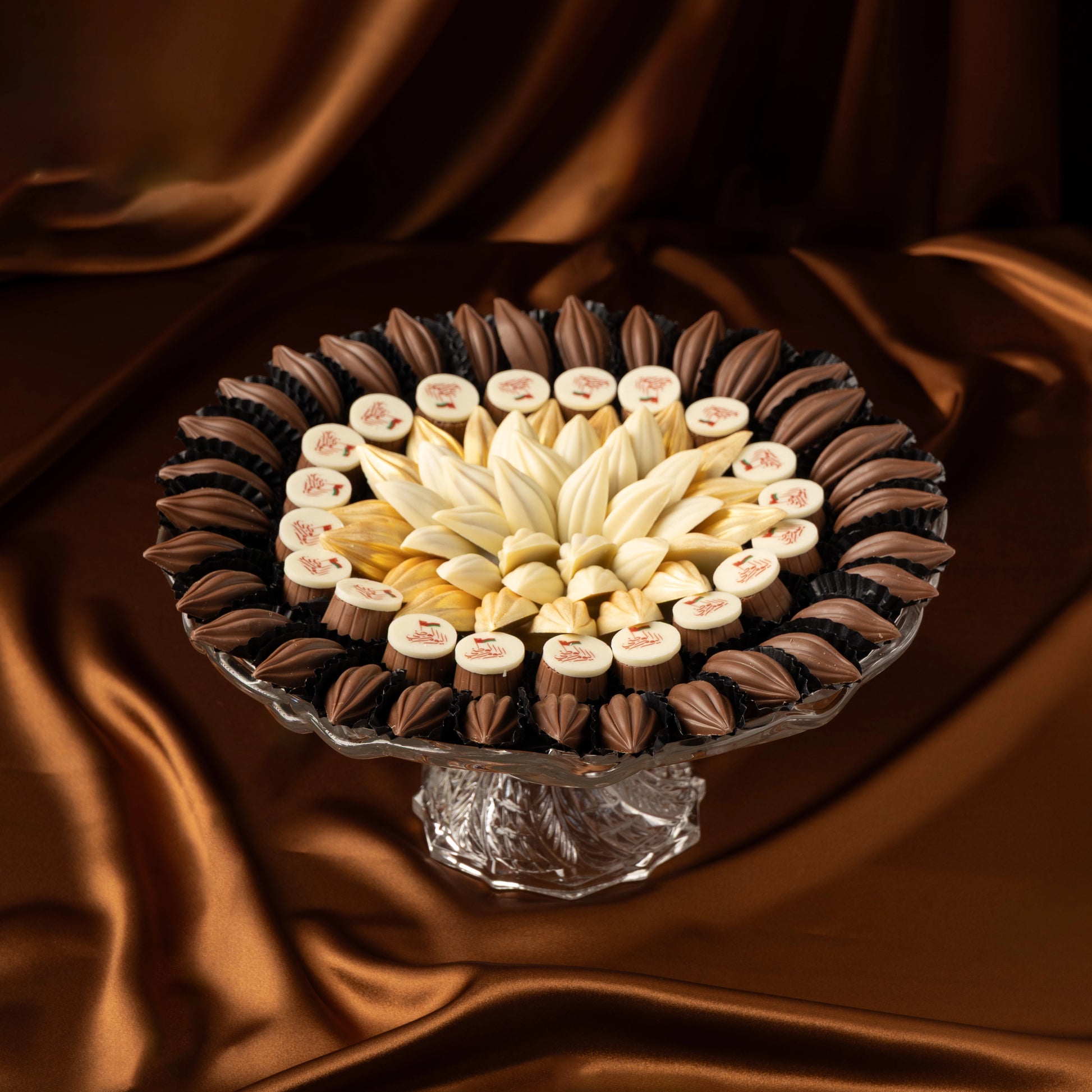 Decorative arrangement of Omani National Day chocolate tray with white chocolate and milk chocolate on a glass stand against a brown fabric background